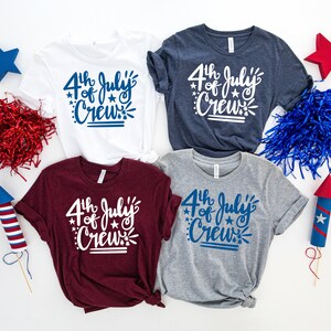 May include: Four t-shirts in white, navy, burgundy, and gray, each with the text "4th of July Crew" in a script font. Decorative stars and fireworks elements surround the text. Patriotic theme.