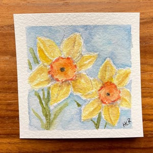 May include: Watercolor painting of two yellow daffodils with orange centers and green stems against a light blue background. The artwork is on a square piece of white paper, signed in the bottom right corner.