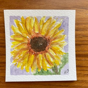 May include: A square watercolor painting on textured paper featuring a single sunflower with bright yellow petals and a dark brown center. The flower is set against a light purple background with green leaves at the base and the initials HLR in the corner.