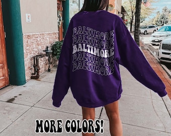 May include: Purple sweatshirt with the word "Baltimore" repeated in a retro font on the back. The text is white and the sweatshirt is oversized. The model is wearing white sneakers and white socks.