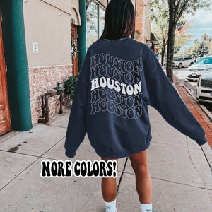 May include: A navy blue sweatshirt with the word "HOUSTON" repeated in a retro, wavy font on the back. The text is white. The sweatshirt is being worn by a person walking down a sidewalk. The text "MORE COLORS!" is printed in white on the sidewalk.
