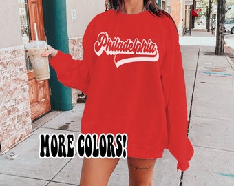 May include: Red sweatshirt with white text that says "Philadelphia" in a retro font. The text is in a curved shape, like a baseball logo. The sweatshirt is being worn by a person with a coffee cup in their hand. The text "MORE COLORS!" is at the bottom of the image.