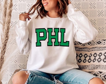 Puede incluir: Sudadera blanca con letras verdes que dicen "PHL" en la parte delantera. La sudadera la lleva puesta una persona sentada en un sofá.