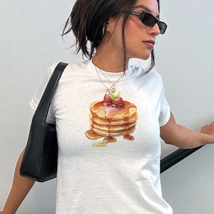 May include: A white t-shirt with a graphic of a stack of pancakes topped with strawberries and syrup. The person wearing the shirt is also wearing a black floral skirt and sunglasses.