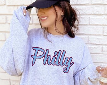 May include: A gray sweatshirt with the word "Philly" in blue and maroon lettering. The sweatshirt is being worn by a person with a black baseball cap.