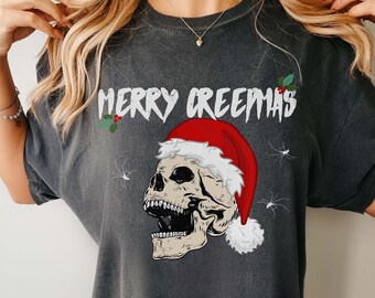 May include: Dark gray t-shirt with the text "MERRY CREEPMAS" in white, featuring a skull wearing a Santa hat. The design includes holly and spider graphics, creating a spooky holiday theme.