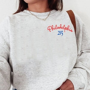 May include: A gray sweatshirt with a red and blue embroidered design that says "Philadelphia 215" on the left chest.