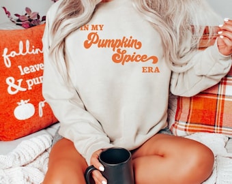 May include: A beige sweatshirt with the text "IN MY Pumpkin Spice ERA" in orange lettering. The person is holding a black mug. Orange and white pillows are in the background.