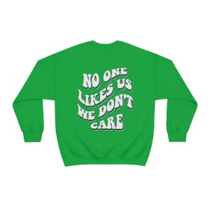 Może przedstawiać: Zielony sweter z białym napisem "No one likes us we don't care" w retro czcionce.