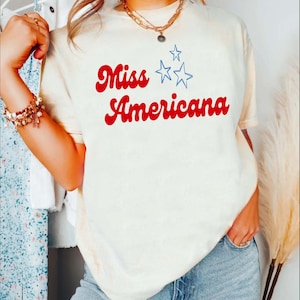 Puede incluir: Una camiseta blanca con el texto "Miss Americana" en rojo con estrellas azules.