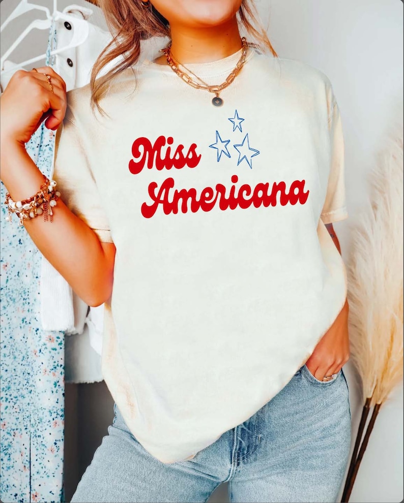 Puede incluir: Una camiseta blanca con el texto "Miss Americana" en rojo con estrellas azules.