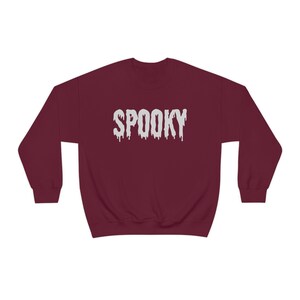 May include: A maroon sweatshirt with the word "SPOOKY" in dripping white letters across the chest. The sweatshirt is a crew neck style, perfect for Halloween or spooky season.