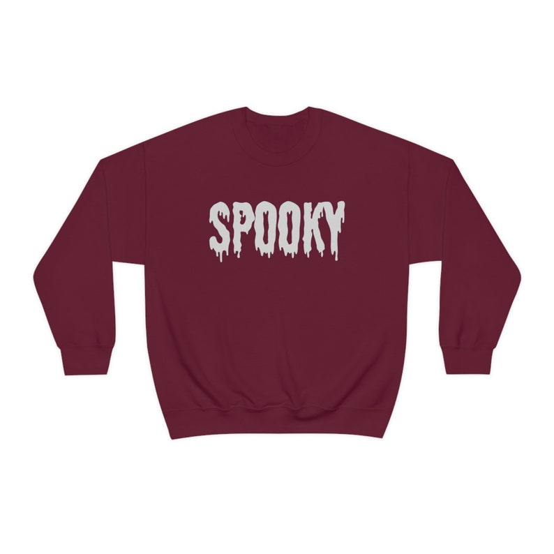 May include: A maroon sweatshirt with the word "SPOOKY" in dripping white letters across the chest. The sweatshirt is a crew neck style, perfect for Halloween or spooky season.