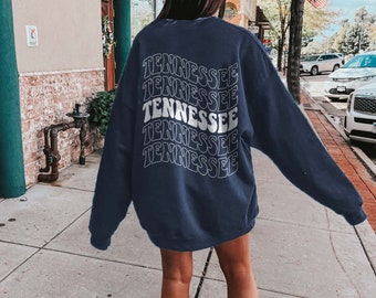 May include: A navy blue sweatshirt with the word "Tennessee" repeated five times in a white retro font on the back. The sweatshirt is worn by a person standing on a sidewalk in front of a building.