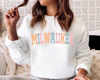 Może przedstawiać: Biała bluza z napisem "MILWAUKEE" na piersi w pastelowych kolorach. Litery są pomarańczowe, niebieskie i różowe. Bluza ma długie rękawy i okrągły dekolt.