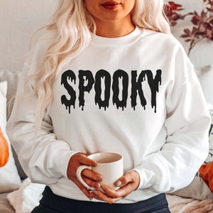 May include: White sweatshirt with the word "SPOOKY" in dripping black letters. The sweatshirt is part of a Halloween-themed collection. The image also includes a pumpkin pillow and a mug.