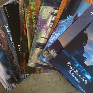 May include: A collection of ten brochures featuring scenic photographs of waterfalls in Arkansas. The brochures are arranged in a fan shape, with the top brochure showing a night sky with stars and the bottom brochure showing a waterfall in a forest.