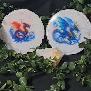 Op de afbeelding: Een set van vier ronde witte onderzetters met achthoekige randen, elk voorzien van een unieke, kleurrijke drakenillustratie in blauwe, oranje en rode tinten. De onderzetters hebben een gespikkelde afwerking en zijn omringd door groen blad.