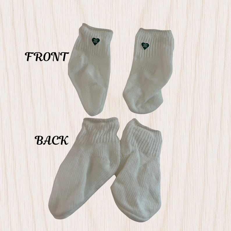 May include: Four white ankle socks, two showing the front and two showing the back. Each sock has a small green heart-shaped emblem near the top. The word "FRONT" and "BACK" are printed in black on the image.