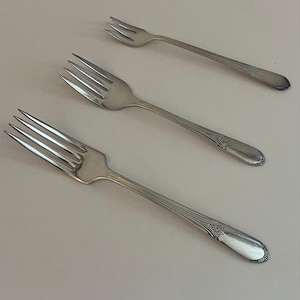 May include: Three silver forks with tines and decorative handles are arranged diagonally on a light-colored surface. The forks have a classic design, suitable for formal dining or special occasions. The handles feature an ornate pattern.