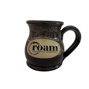 May include: A dark brown ceramic mug with a glossy finish and a flared rim. The front features an oval cream-colored emblem with the black text "roam" inside a circular brushstroke design, followed by the words "INNOVATIVE WORKPLACE" below it.