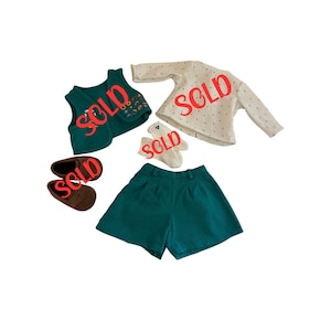May include: A children's clothing set featuring a teal vest with floral embroidery, a cream-colored long-sleeve shirt with small black dots, teal shorts, brown shoes, and white socks. The word "SOLD" is stamped across each item.