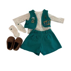 May include: A child's outfit set featuring a teal vest with patches, matching shorts, a long-sleeved shirt with small white dots, white socks, and brown loafers. The vest has a rainbow and the number 101 on it. The outfit is displayed on a white background.