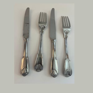 May include: Four pieces of silverware, including two knives and two forks, are arranged on a white surface. The silverware is silver-colored with a decorative shell design on the handles. The knives have a smooth, flat blade.