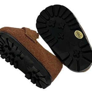 May include: Brown suede-like shoes with black lug soles. The shoes have a classic design with a textured upper and a sturdy, durable sole. The soles have a pattern for traction. The shoes are likely for children.