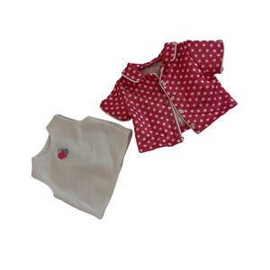May include: Two-piece doll clothing set. A white sleeveless top with a small floral design and a red polka dot pajama top with white trim and buttons. The set is designed for a doll.