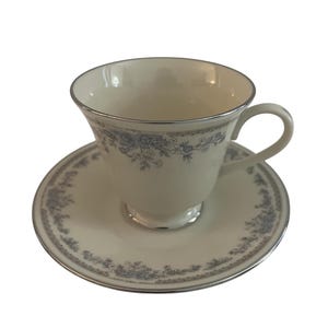 Könnte beinhalten: Ein weißes Tassen- und Untertassen-Set mit zartem blauem Blumenmuster und silberner Verzierung. Die Tasse hat einen gebogenen Henkel und steht auf einer passenden Untertasse. Das Set ist wahrscheinlich aus Porzellan.