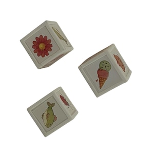 May include: Three white toy blocks with illustrations of a red flower, a fish, and an ice cream cone. The blocks are slightly angled, showcasing different sides with various drawings. The illustrations are in soft, muted colors.