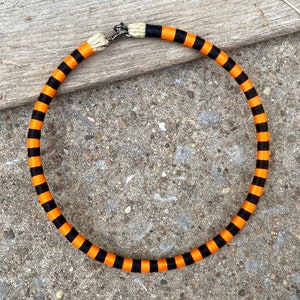 Puede incluir: Un collar de rayas negras y naranjas con un cierre plateado. El collar est&aacute; hecho de tela y mide aproximadamente 45 cm de largo.