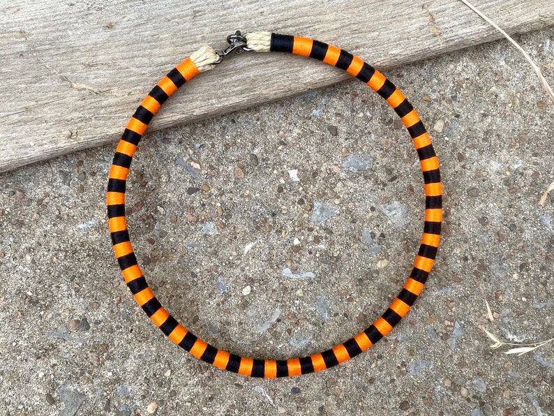 Puede incluir: Un collar de rayas negras y naranjas con un cierre plateado. El collar est&aacute; hecho de tela y mide aproximadamente 45 cm de largo.