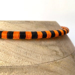 Puede incluir: Un cord&oacute;n rayado de color negro y naranja, posiblemente para un collar o una pulsera, est&aacute; sobre una superficie de madera.