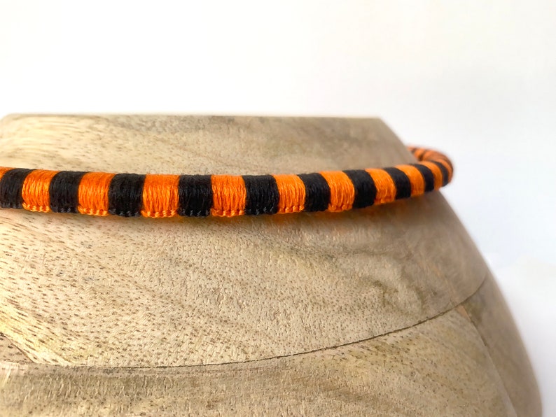Puede incluir: Un cord&oacute;n rayado de color negro y naranja, posiblemente para un collar o una pulsera, est&aacute; sobre una superficie de madera.