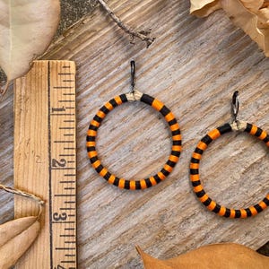 Peut inclure: Une paire de boucles d'oreilles cr&eacute;oles ray&eacute;es orange et noires. Les boucles d'oreilles sont en tissu et ont un crochet en m&eacute;tal noir.