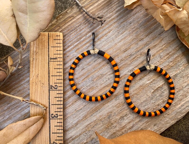 Peut inclure: Une paire de boucles d'oreilles cr&eacute;oles ray&eacute;es orange et noires. Les boucles d'oreilles sont en tissu et ont un crochet en m&eacute;tal noir.