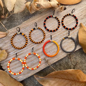 Peut inclure: Un ensemble de six paires de boucles d'oreilles cr&eacute;oles, chacune avec une combinaison de couleurs diff&eacute;rente de noir, orange et blanc. Les boucles d'oreilles sont pr&eacute;sent&eacute;es sur une surface en bois.