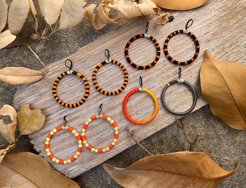Peut inclure: Un ensemble de six paires de boucles d'oreilles cr&eacute;oles, chacune avec une combinaison de couleurs diff&eacute;rente de noir, orange et blanc. Les boucles d'oreilles sont pr&eacute;sent&eacute;es sur une surface en bois.