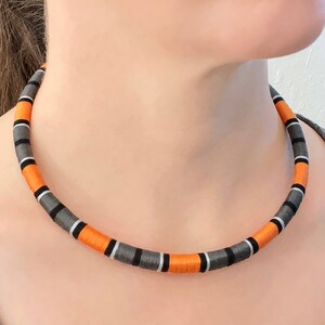 May include: A close-up of a necklace made of orange, gray, and black woven fabric. The necklace is worn by a person, but the person's face is not visible.