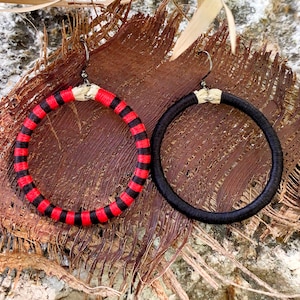 Puede incluir: Un par de pendientes de aro, uno negro y otro con rayas negras y rojas. Los pendientes están hechos de tela y cuelgan de un fondo natural.