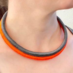 Può includere: Un primo piano di una collana choker in tessuto grigio e arancione. La collana è composta da due fili di tessuto, uno grigio e uno arancione, che sono intrecciati insieme.