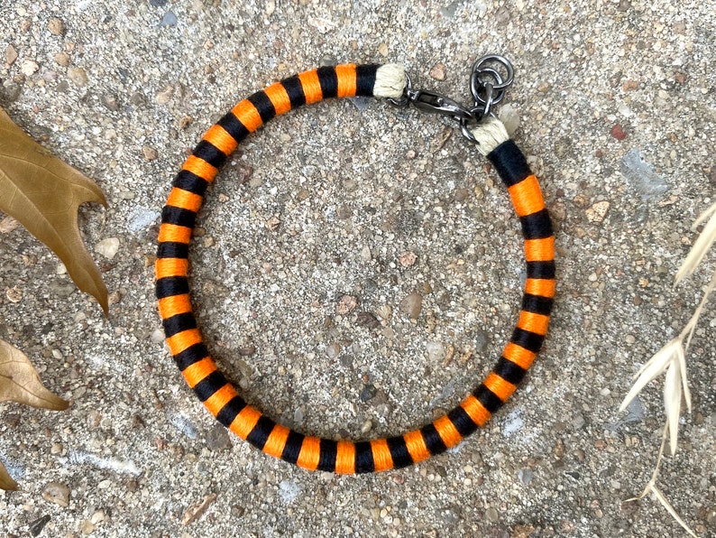 Peut inclure: Un collier en corde ray&eacute; noir et orange avec une boucle en m&eacute;tal argent&eacute;.