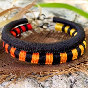 Puede incluir: Dos pulseras de tela negra con rayas naranjas y amarillas. Las pulseras están apiladas una encima de la otra.