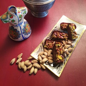 Op de afbeelding: Een zilveren kom met een deksel, een keramische vaas met een bloemenpatroon en een bord met pistache- en chocoladevierkanten. Het bord heeft een zwart-wit zebrapatroon met een gouden rand.