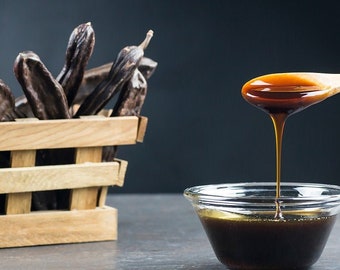 Puede incluir: Una cuchara de madera vierte jarabe de algarroba marrón oscuro en un tazón de vidrio transparente. En el fondo hay una cesta de vainas de algarroba secas.