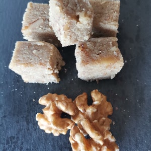 Puede incluir: Primer plano de un grupo de cinco dulces cuadrados, masticables, de color marr&oacute;n claro, rellenos de nueces, sobre una superficie de pizarra negra. Tres nueces enteras se encuentran en la esquina inferior derecha de la imagen.