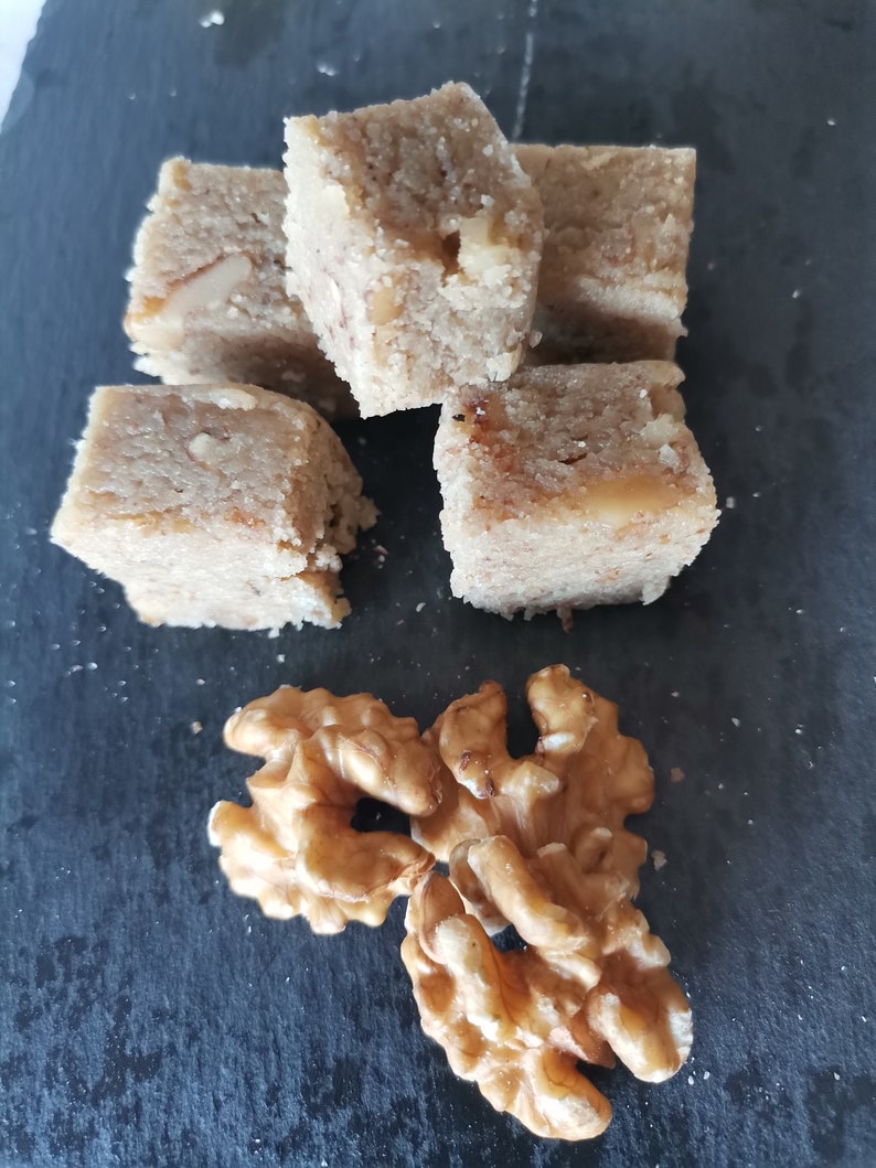 Puede incluir: Primer plano de un grupo de cinco dulces cuadrados, masticables, de color marr&oacute;n claro, rellenos de nueces, sobre una superficie de pizarra negra. Tres nueces enteras se encuentran en la esquina inferior derecha de la imagen.