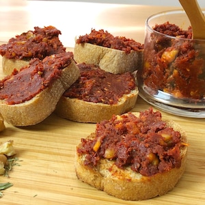 May include: A close-up of a wooden cutting board with several slices of bread topped with a red spread. The spread is in a glass jar with a wooden spoon. There are also walnuts and herbs on the board.